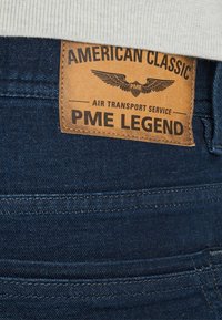 Tamno plavi traperice s smešnim kožnim oznakom koja sadrži "PME LEGEND" i dizajn krila. Oznaka je ušivena na stražnjem pojasu.