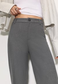 Sive široke hlače ravnog kroja s glatkom teksturom, s prednjim zatvaranjem na kopče, bočnim džepovima i krojem po mjeri. Nošene s laganim gornjim dijelom.