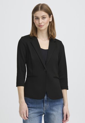 Noor naine, kes kannab musta kolmnurgaga bleiserit, millel on kolmveerandvarrukad, musta pluusi ja siniseid teksaseid, seisab tavalise taustaga seina ees.