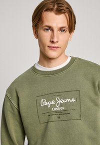 Sudadera verde de algodón, con un parche de logo blanco que lleva el texto "Pepe Jeans London". Cuello redondo y textura suave.