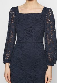 Navy blue lace dress featuring a square neckline, long sheer sleeves, floral patterns, and a fitted waist with delicate embroidery accents.