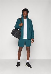 Turkos lätt jacka med huva, vit t-shirt, turkosa shorts, svarta sneakers med gula accenter och en svart duffelväska.