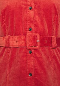 Vêtement en velours côtelé rouge avec des boutons noirs au centre et une ceinture assortie texturée avec une boucle carrée à la taille.