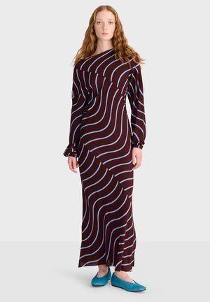 Femme aux longs cheveux roux portant une longue robe sombre avec des rayures ondulées bleu clair et des chaussures plates bleu vif, debout devant un fond uni.