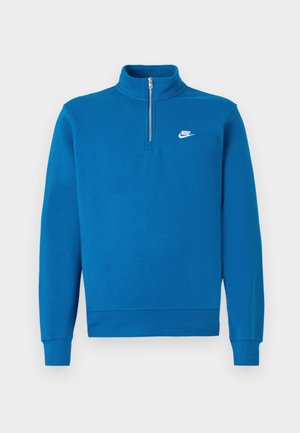 Zils Nike džemperis ar augstu apkakli, pus-izjūgu priekšpusi un balto logotipu kreisajā krūtī; izgatavots no mīksta, teksturēta auduma.