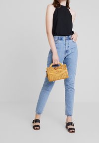Sac à main jaune tissé avec fermeture à bouton en bois, haut noir sans manches, jean taille haute bleu clair, et sandales plates noires.