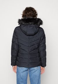 Schwarze Daunenjacke mit einer Kapuze aus Kunstfell, mit einem Chevron-Steppmuster, Reißverschluss-Taschen und elastischen Bündchen. Rückansicht angezeigt.