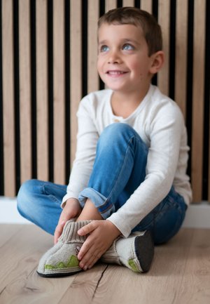 Junger Junge in weißem Hemd und blauer Jeans sitzt auf dem Boden, lächelt und richtet graue Wollhausschuhe mit grünen Akzenten.