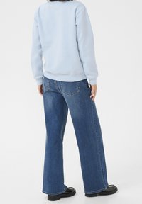 Felpa azzurra con polsini e orlo a costine, abbinata a jeans a gamba larga blu scuro con tasche posteriori e cuciture visibili.