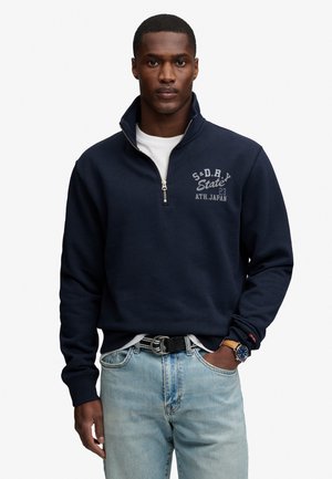 Hombre con sudadera azul marino con cremallera hasta la mitad y texto en el pecho, vaqueros azul claro, cinturón negro y reloj de pulsera, de pie sobre fondo blanco.