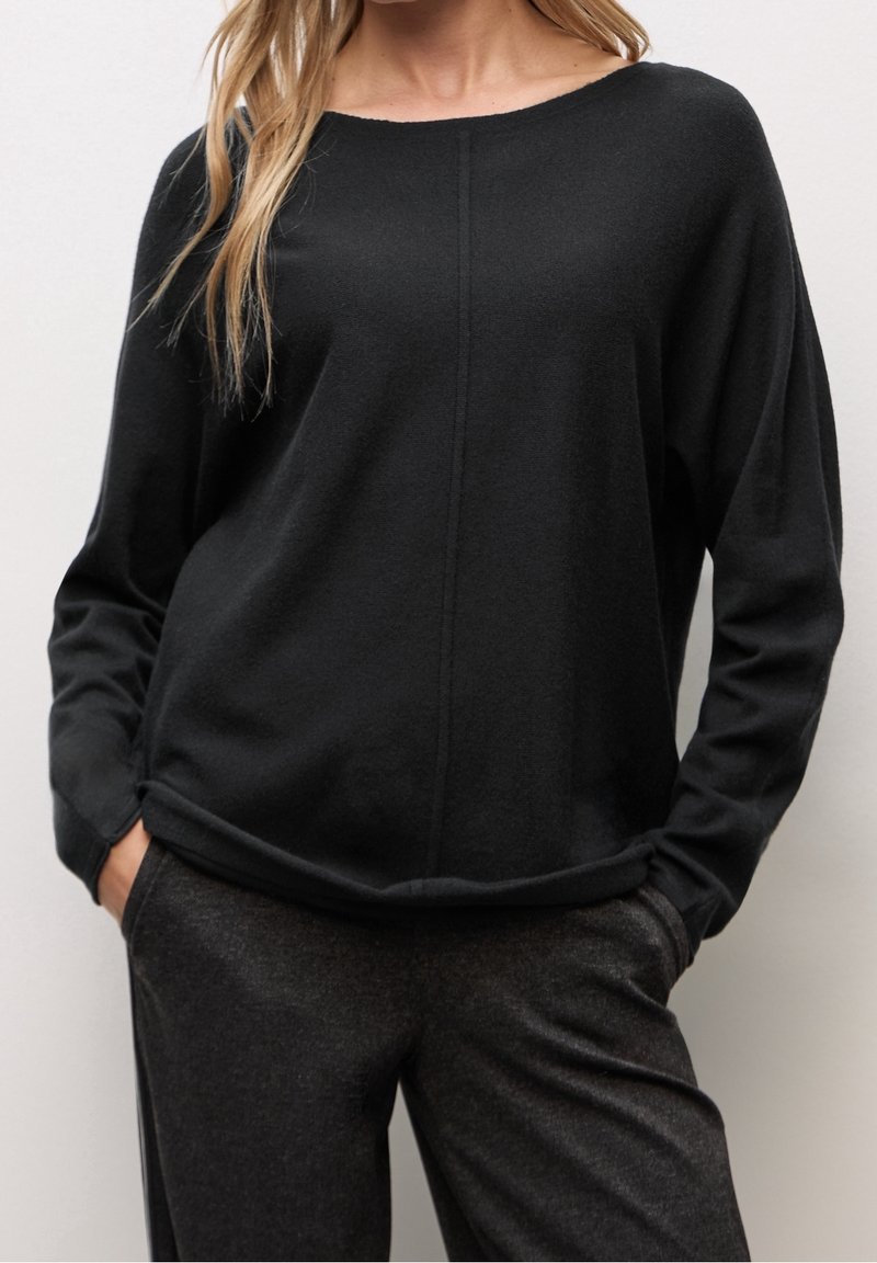 Femme portant un haut noir ample à manches longues avec une couture centrale et un pantalon gris foncé, mains dans les poches devant un fond uni.