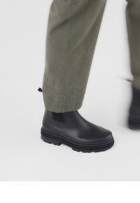 Crne gumene gležnjače s elastičnim bočnim panelima, teksturiranim potplatom i zaobljenim vrhom, uparene s maslinasto zelenim hlačama.