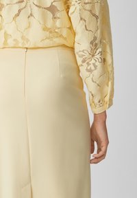 Ljusgul blus med blommig spetsmönster och långa ärmar, bärs över matchande ljusgula byxor med en slät textur och skräddarsydd passform.