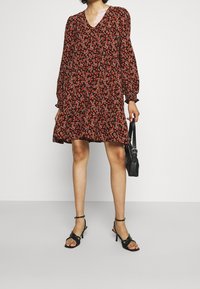 Vestito floreale nero con maniche a sbuffo lunghe, scollo a V e orlo a più strati. Abbinato a sandali neri con tacco e una piccola borsa a tracolla.
