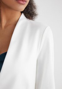 Witte blazer met een gladde, matte stofafwerking. Heeft een notch-kraag, een getailleerde pasvorm en subtiele stiksels langs de randen.