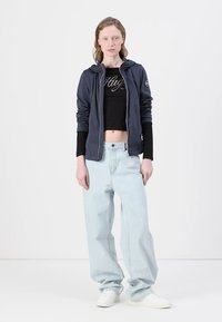 Mornarsko zip up kapuco čez črn cropped top z besedilom "Hug", v kombinaciji s svetlo modrimi širokimi hlačami in belimi supergami.