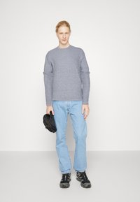 Maglione grigio a maniche lunghe, abbinato a jeans azzurri chiari. Sneakers nere con accenti grigi e una piccola borsa nera tenuta nella mano sinistra.
