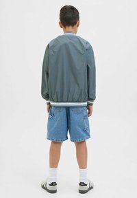 Grön bomberjacka med vita ribbade muddar och krage, bärs med blå denimshorts. Bakifrån syns det elastiska midjebandet och två sidofickor.