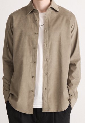 Man draagt een beige losse lange mouwen overhemd over een wit T-shirt met zwarte broek, handen in de zakken, tegen een effen achtergrond.