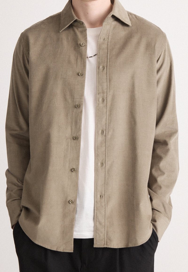 Homme portant une chemise beige à manches longues déboutonnée sur un t-shirt blanc avec un pantalon noir, les mains dans les poches, sur un fond uni.