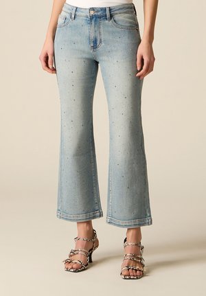 Frau trägt hellblaue ausgestellte Jeans mit kleinen Strassdetails und High-Heels mit Schlangenprint, steht vor einem schlichten Hintergrund.