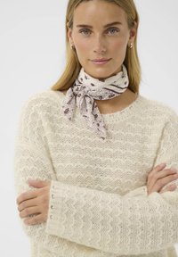 Pull en tricot crème avec un motif de vagues texturé, manches longues et poignets côtelés. Porté avec une écharpe légère à motif nouée autour du cou.