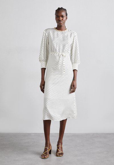 Polka dot midi dress in white fabric with black dots, featuring puffed sleeves, a cinched waist, and a tie detail. Worn with strappy heels.
