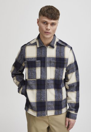 Veste à carreaux avec de grands motifs navy, crème et beige, dotée d'une fermeture zippée, d'un col et de deux poches avant. Matériau texturé ressemblant à de la laine.