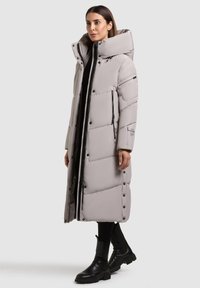 Kvinde iført lang lys grå dunfrakke med høj krave, sorte leggings og sorte ankelstøvler, stående mod en grå baggrund.