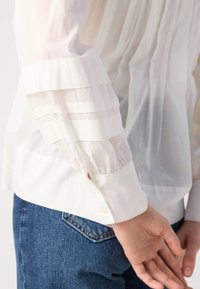 White blouse featuring sheer, layered sleeves with elastic cuffs, complemented by a buttoned wrist, paired with blue denim jeans.