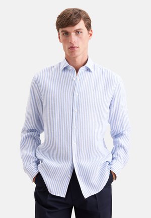 Ung mand iført en lyseblå og hvid lodretstribet skjorte med knapper og lange ærmer, med hænderne i de mørke bukselommer.