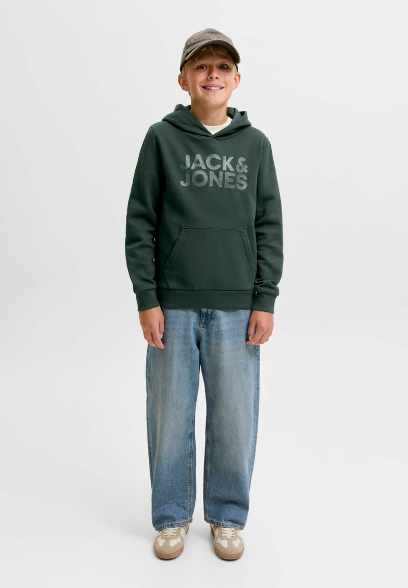 Αγόρι που φοράει πράσινο hoodie Jack & Jones, φαρδιά ανοιχτά μπλε τζιν, μπεζ αθλητικά παπούτσια και γκρι καπέλο, στέκεται και χαμογελάει σε λευκό φόντο.