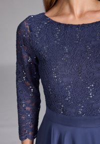Robe bleu marine avec un bustier en dentelle ornée de sequins, longues manches transparentes et jupe lisse. Motif floral texturé sur le dessus.