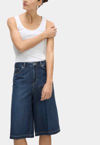 Dunkelblaue Denim-Culottes mit einem weiten Bein-Design. Verfügt über vordere Taschen, einen Knopfverschluss und sichtbare Nahtakzente. Kombiniert mit einem weißen Tanktop.