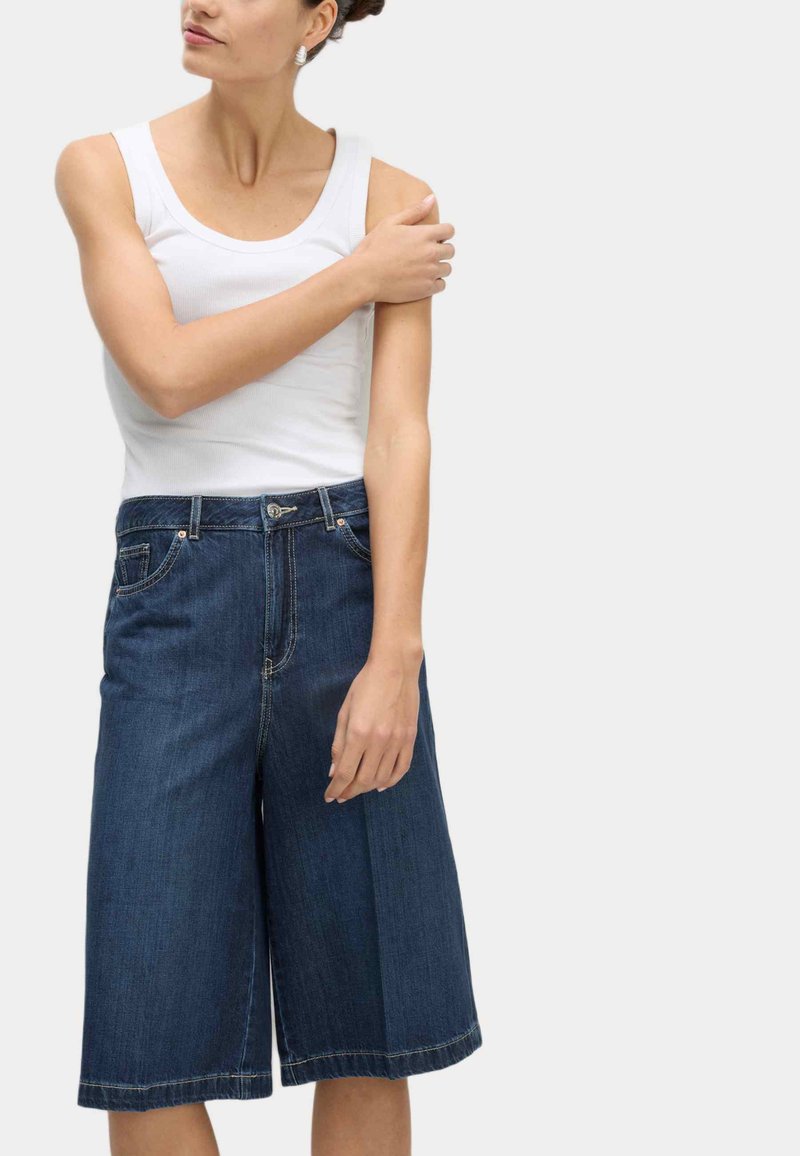 Dunkelblaue Denim-Culottes mit einem weiten Bein-Design. Verfügt über vordere Taschen, einen Knopfverschluss und sichtbare Nahtakzente. Kombiniert mit einem weißen Tanktop.
