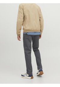 Beige Bomberjacke, graue Hose und blaue Sneakers mit Gummisohle. Die Jacke hat einen gerippten Saum und elastische Bündchen. Sichtbares Jeanshemd darunter.