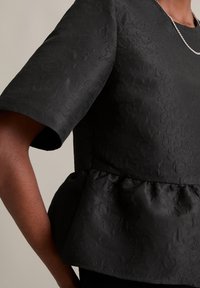 Melnā pepluma blūze ar īsām piedurknēm, kas raksturojas ar faktūru. Gluda auduma ar vieglu spīdumu. Mazs ķēdes kaklarota kā akcents.