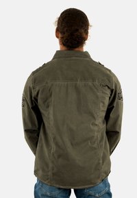 Giacca militare verde oliva, con spalline, maniche lunghe e taglio rilassato. Presenta una texture liscia e dettagli cuciti.