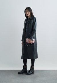 Manteau trench en cuir noir avec des boutons, associé à des leggings noirs et des bottes de combat. Tenant un petit sac à main rose. Design simple et élégant.