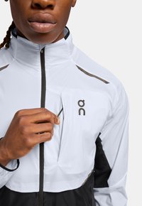 Hvid løbejacket med sorte accenter, der har høj krave, lynlås foran og en brystlomme. Letvægtigt stof og slankt design.