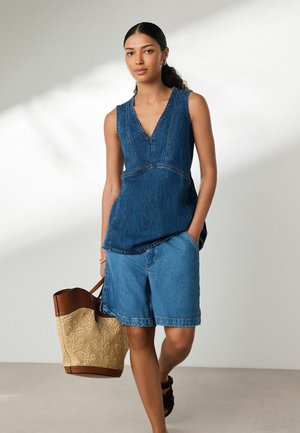 Femeie care poartă un top din denim fără mâneci și pantaloni scurți, ținând o geantă mare țesută cu accente din piele.