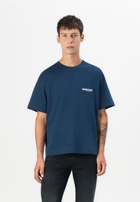 Navy blue cotton t-shirt with short sleeves, crew neckline, and a small white logo on the left chest. Textured fabric with a relaxed fit.