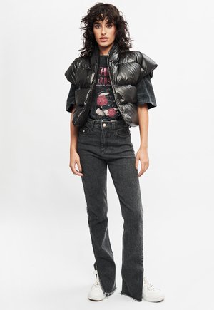 Kobieta z kręconymi ciemnymi włosami, ubrana w czarną puchową kamizelkę, graficzny t-shirt, czarne dżinsy z wysokim stanem i surowymi brzegami oraz białe trampki, stojąca na tle białego tła.