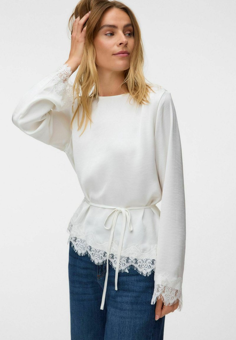 Femme aux longs cheveux blonds portant un chemisier blanc à manches longues avec bordure en dentelle et taille nouée, associé à un jean bleu, touchant ses cheveux.