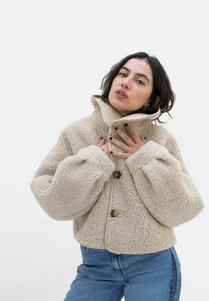Beige faux fur jakke med høj krave, store lommer og træknapper. Blødt, struktureret stof, cropped design. Sammen med blå jeans.