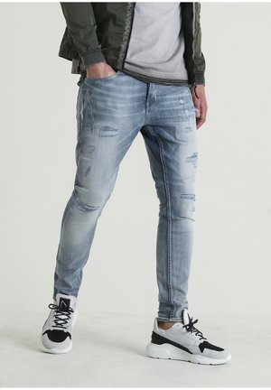 Personne portant un jean skinny bleu clair déchiré et des baskets blanches avec des accents noirs, une main dans la poche et une veste décontractée.