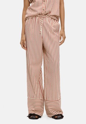 Luźne kremowe spodnie w pionowe czerwone paski, szerokie nogawki i mankiet z poziomym paskiem, noszone z czarnymi sandałami z otwartymi palcami.