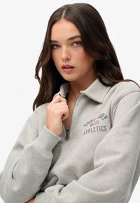 Sudadera de color gris claro con media cremallera y puños acanalados, con "Superdry Athletics" en letras oscuras y un acento de la bandera americana.