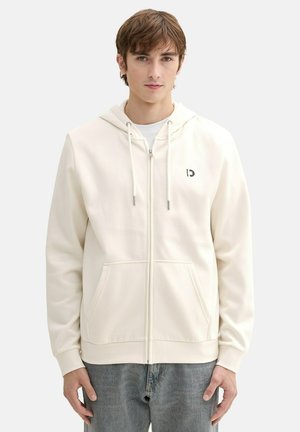 Sudadera cremosa clara con cremallera y capucha ajustable, bolsillos frontales y un pequeño logo en el lado izquierdo del pecho. Tejido suave con una textura lisa.