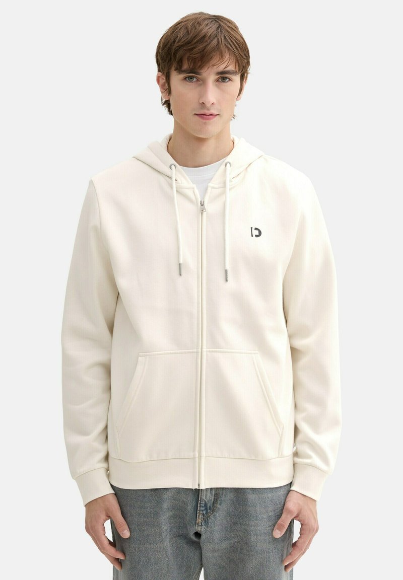 Svetlá krémová mikina na zips s kapucňou na šnúrku, prednými vreckami a malým logom na ľavom hrudníku. Mäkká tkanina s hladkým povrchom.